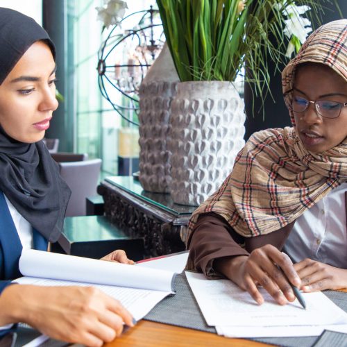 Serious female colleagues studying documents in cafe. Young Muslim business women reading and analyzing papers. Paperwork concept