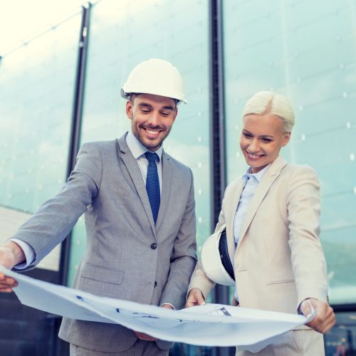 business, partnership, architecture and people concept - smiling businessman and businesswoman with blueprint and helmets on city street